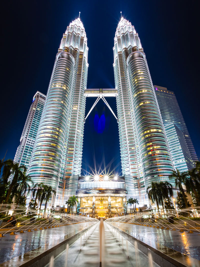 Night view of the Petronas Twin Towers illuminated against the skyline, reflecting over fountains and palm-lined plaza as a proud National Landmark